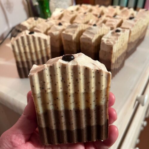 Interior photo of a hand holding a bar of pretty cafe latte soap, with rows of additional bars of handmade soap in the background