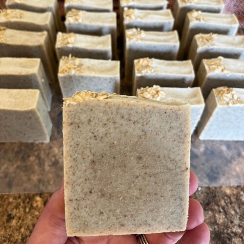 Interior photo of a hand holding a pretty bar of oatmeal chamomile soap, with rows of other bars of soap in the background