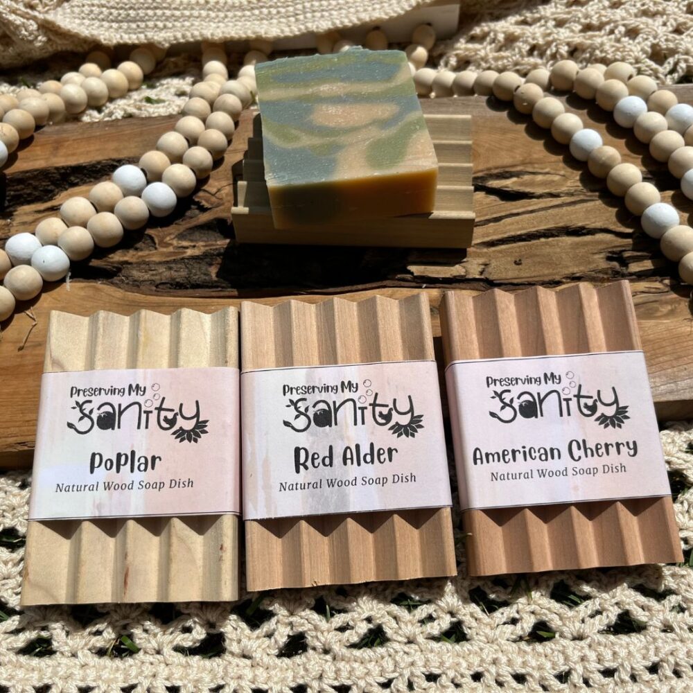 Exterior flatlay photo of a bar of soap on a handmade wood soap holder, with three labeled soap dishes shown in the front of the photo. Backdrop includes rustic wood, wooden beads, and a crocheted sweater Sycamore wood soap dish not pictured.