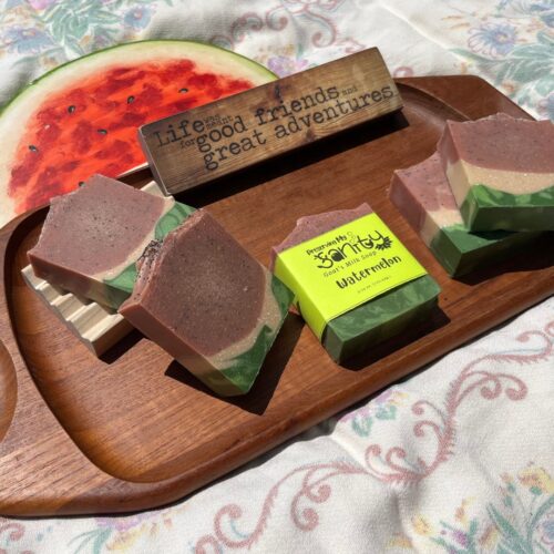 Fun exterior flatlay of watermelon soap with a wooden watermelon on a wooden tray and colorful tablecloth in the sunshine