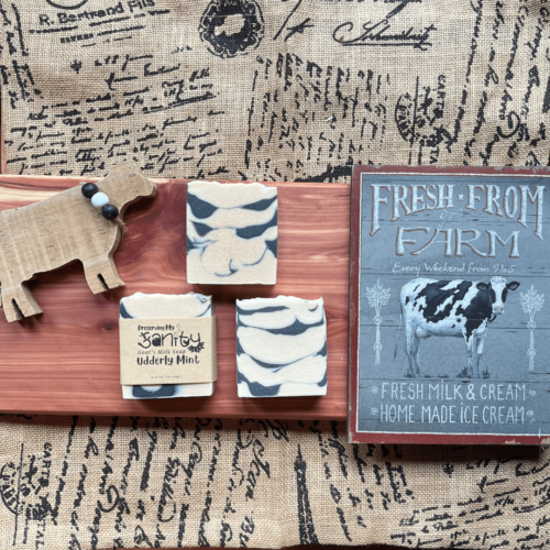Three bars of Udderly Mint essential oil soap with a wooden cow and farm sign on some decorated burlap fabric and a cedar board