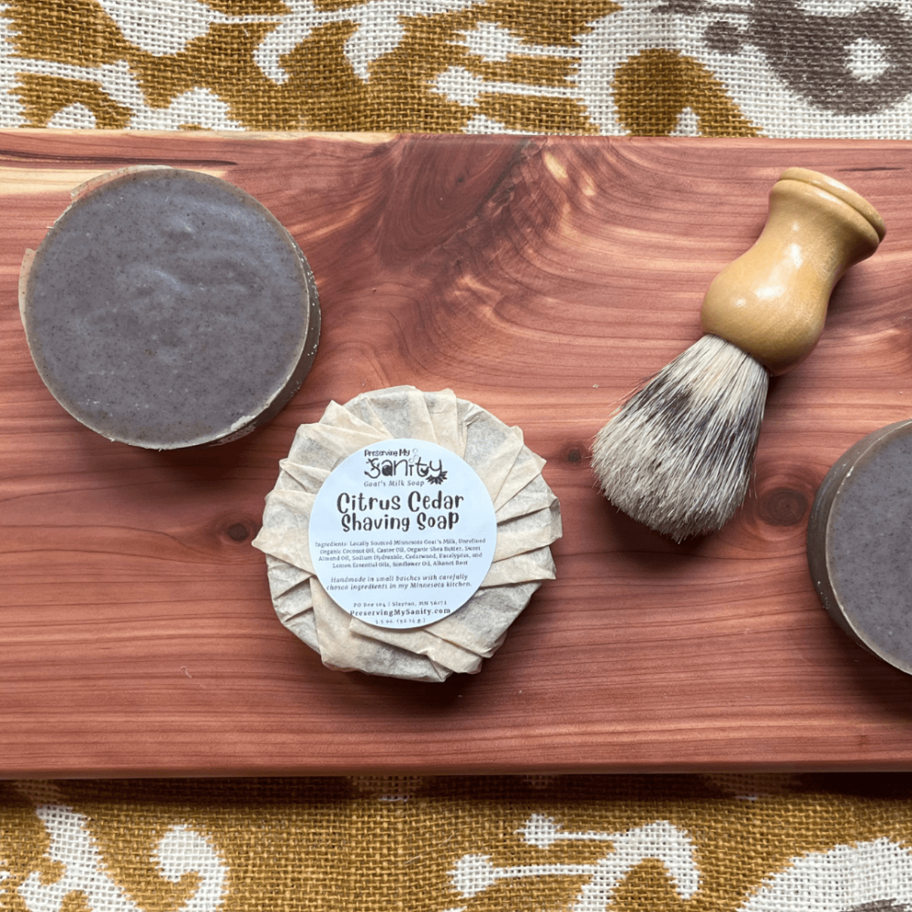 Two round bars of Citrus Cedar Shaving Soap, one wrapped with a label, and a made-in-the-US shaving brush.