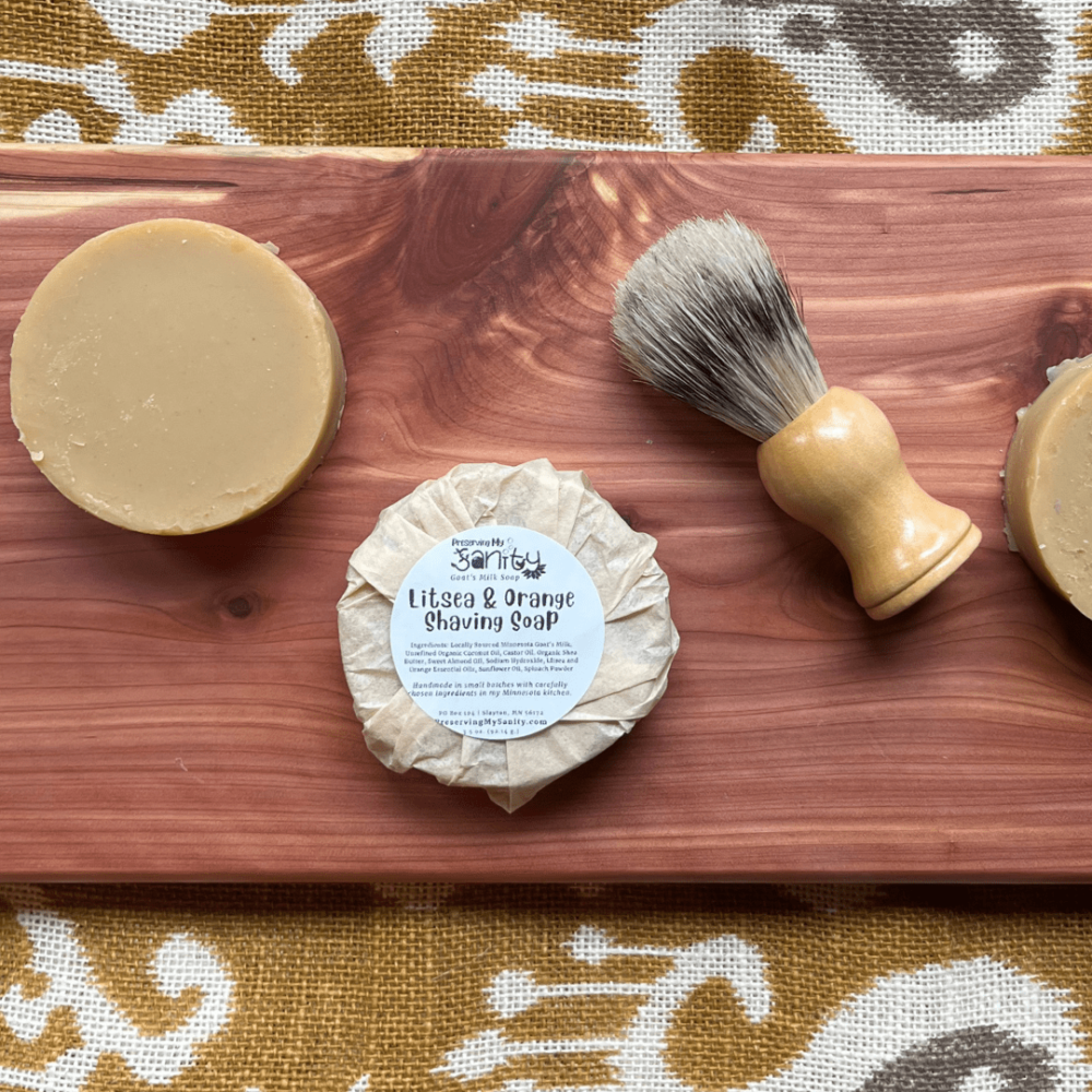 Two round bars of Litsea Orange Shaving Soap, one wrapped with a label, and a made-in-the-US shaving brush.