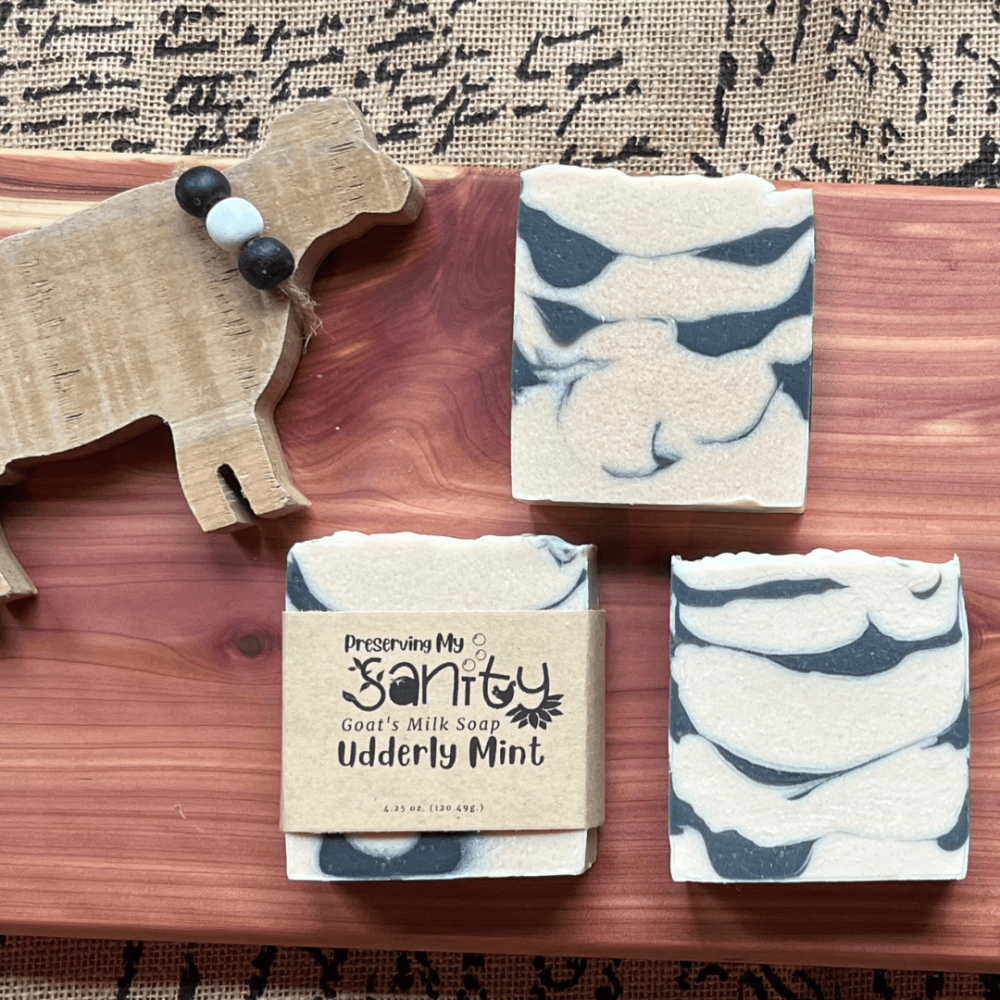 Three bars of Udderly Mint essential oil soap on a cedar board with a wooden cow decoration, with some decorated burlap fabric behind it.