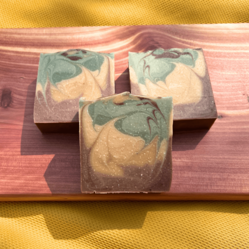 Three bars of Rustic Woods and Rum soap on a cedar board with a dark yellow tablecloth behind it.