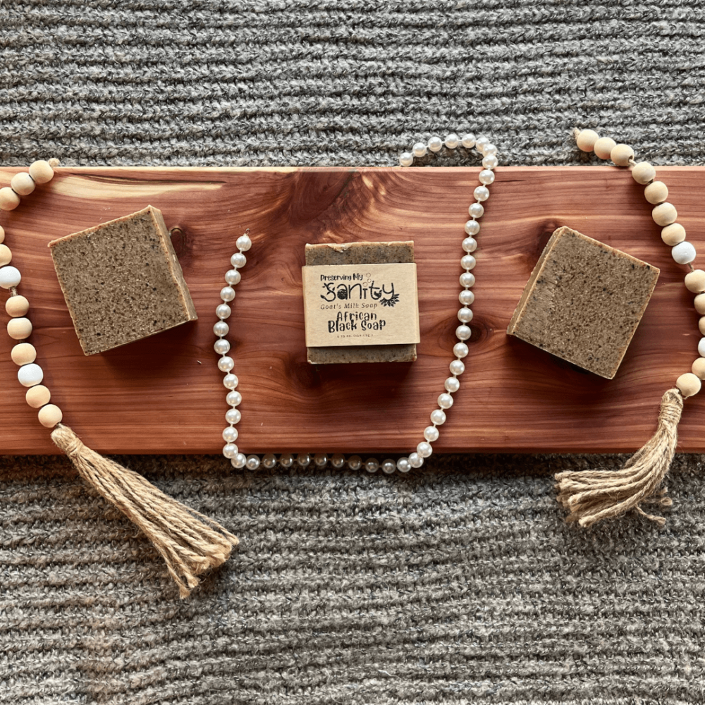 Flatlay of three bars of African Black Soap with goat's milk surrounded by wooden beads, pearls, and a gray sweater on a cedar board.