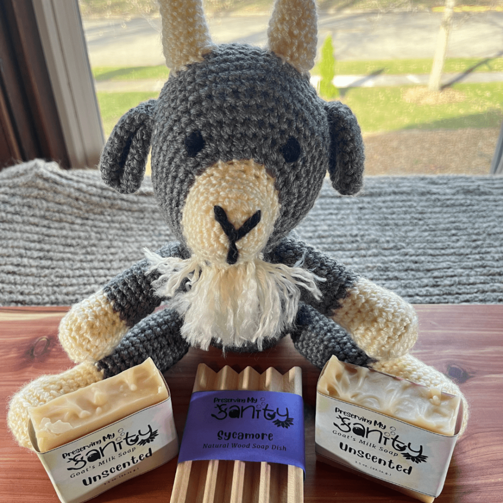 Photo of a handmade goat stuffed animal with two bars of unscented goat's milk soap and a wood soap dish, sitting on a cedar board and gray sweater backdrop.
