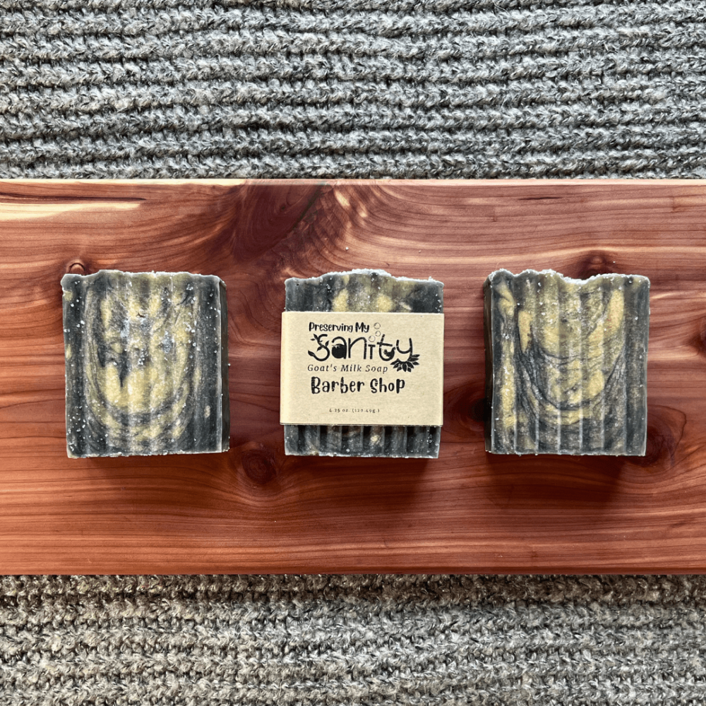 Flatlay photo of three bars of Barber Shop goat's milk soap sitting on a cedar board with a gray sweater backdrop