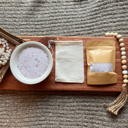 Flatlay photo showing a bowl of lavender bath salt, with a package of bath salt and a reusable cheesecloth bag.