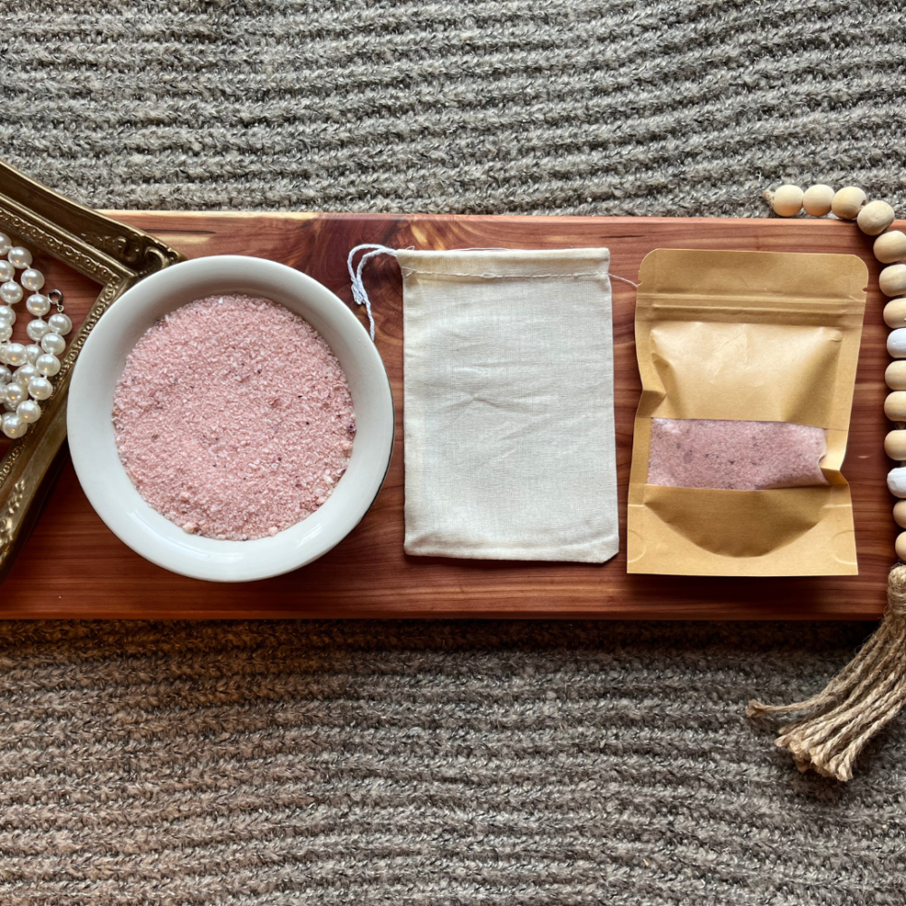 Flatlay photo showing a bowl of rose bath salt, with a package of bath salt and a reusable cheesecloth bag.