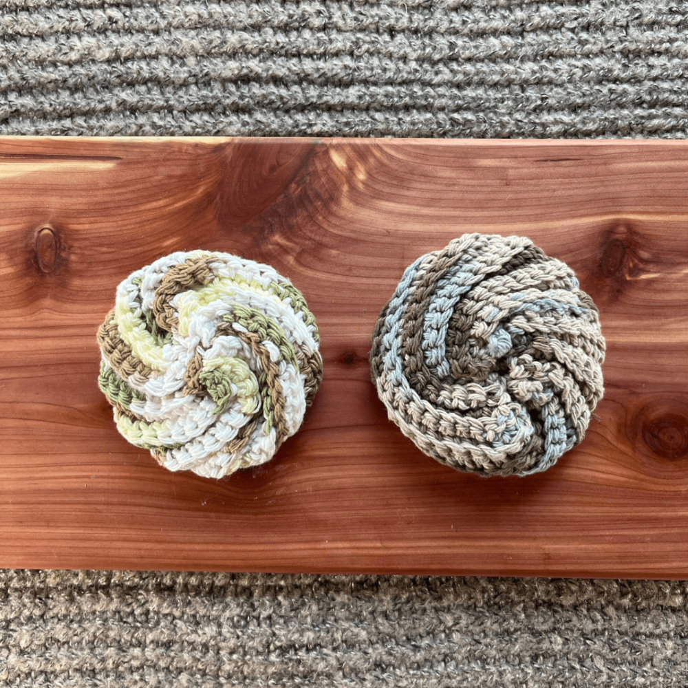 Photo of two different colors of handmade crocheted cotton face scrubbies sitting on a cedar board with a gray sweater backdrop