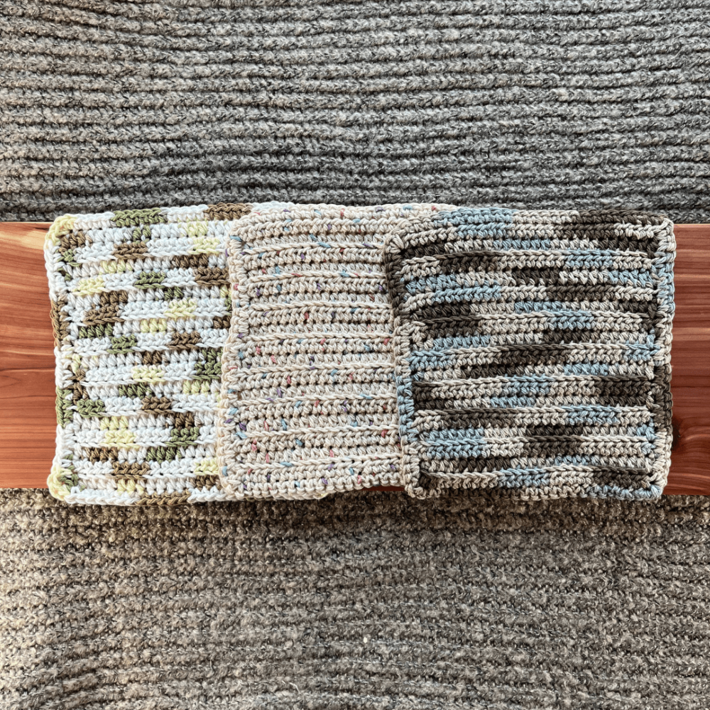 Photo of three different colors of crocheted cotton washcloths sitting on a cedar board with a gray sweater backdrop