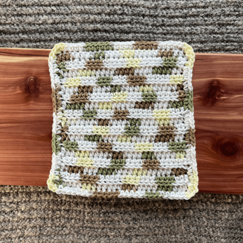 Photo of a variegated white, green, brown, and yellow crocheted cotton washcloth sitting on a cedar board with a gray sweater backdrop