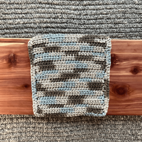 Photo of a variegated tan, brown, and blue crocheted cotton washcloth sitting on a cedar board with a gray sweater backdrop