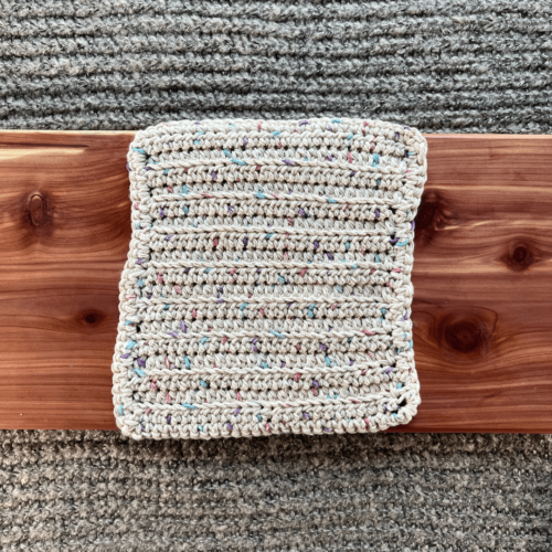 Photo of a variegated beige with pastels crocheted cotton washcloth sitting on a cedar board with a gray sweater backdrop