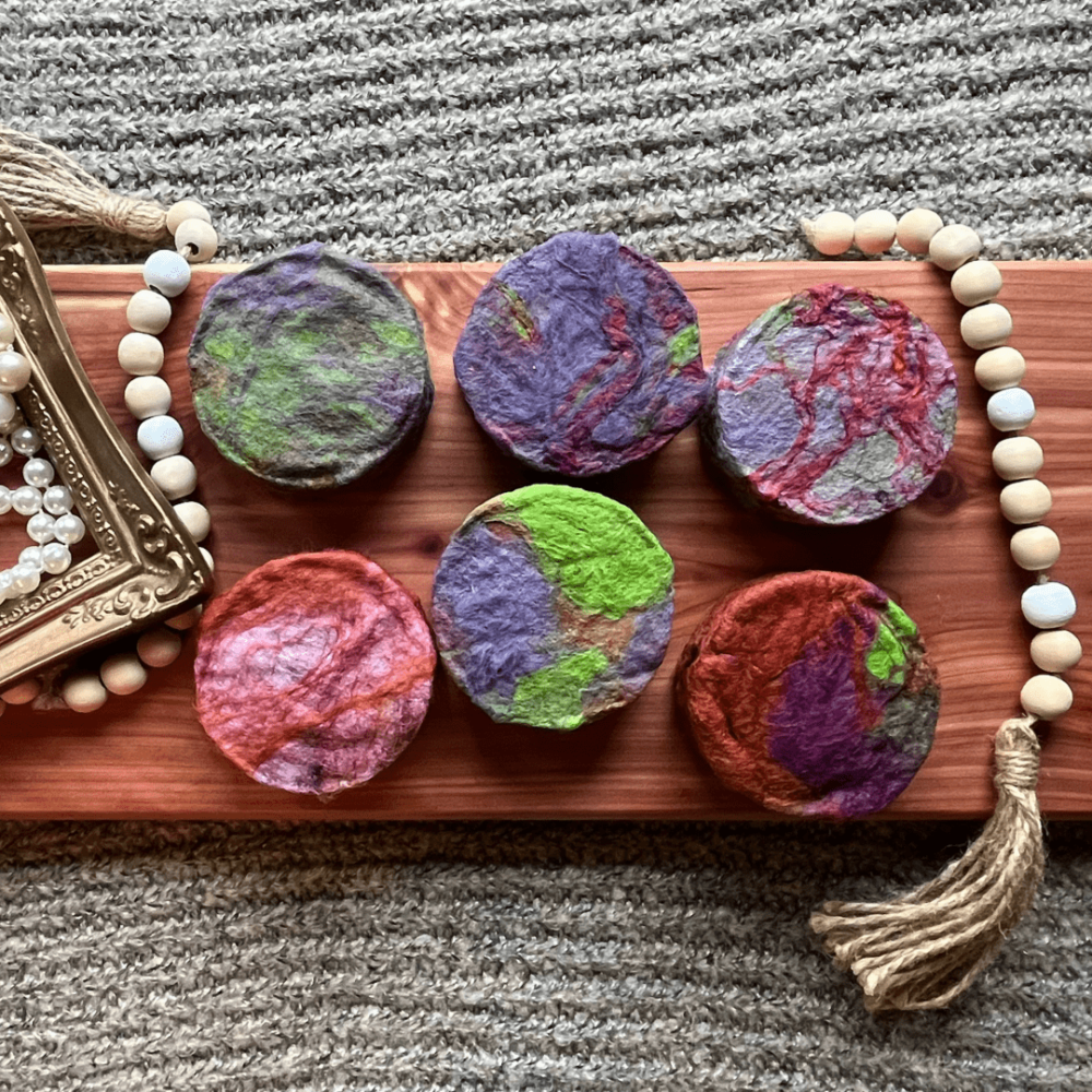 Six bars of colorful felted Nag Champa round bars of soap in greens, blues, and reds on a cedar board
