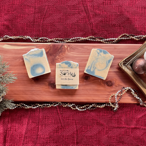 Holiday-themed flatlay of three bars of Fresh Snow goat's milk soap on a cedar board