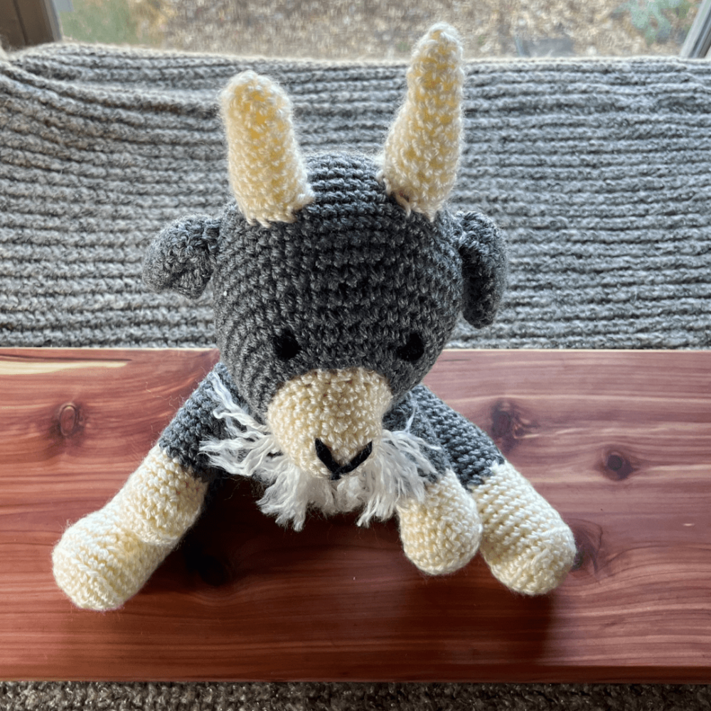 Handmade crocheted goat stuffy sitting on a cedar board with a gray sweater backdrop