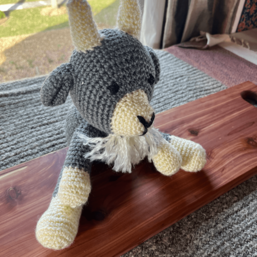 Alternate angle of handmade crocheted goat stuffy sitting on a cedar board with a gray sweater backdrop