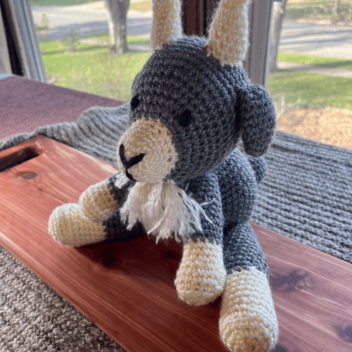 Alternate angle of handmade crocheted goat stuffy sitting on a cedar board with a gray sweater backdrop