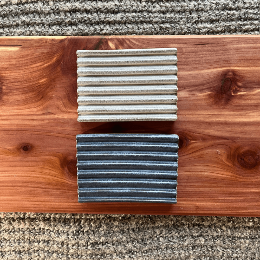 Flatlay photo of two handmade pottery soap dishes on a cedar board with a gray sweater backdrop