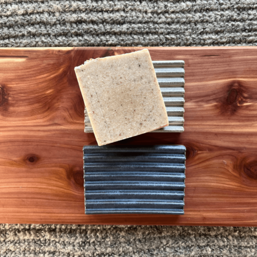 Flatlay photo of two handmade pottery soap dishes on a cedar board with a gray sweater backdrop, with one bar of soap sitting on one of the soap dishes.