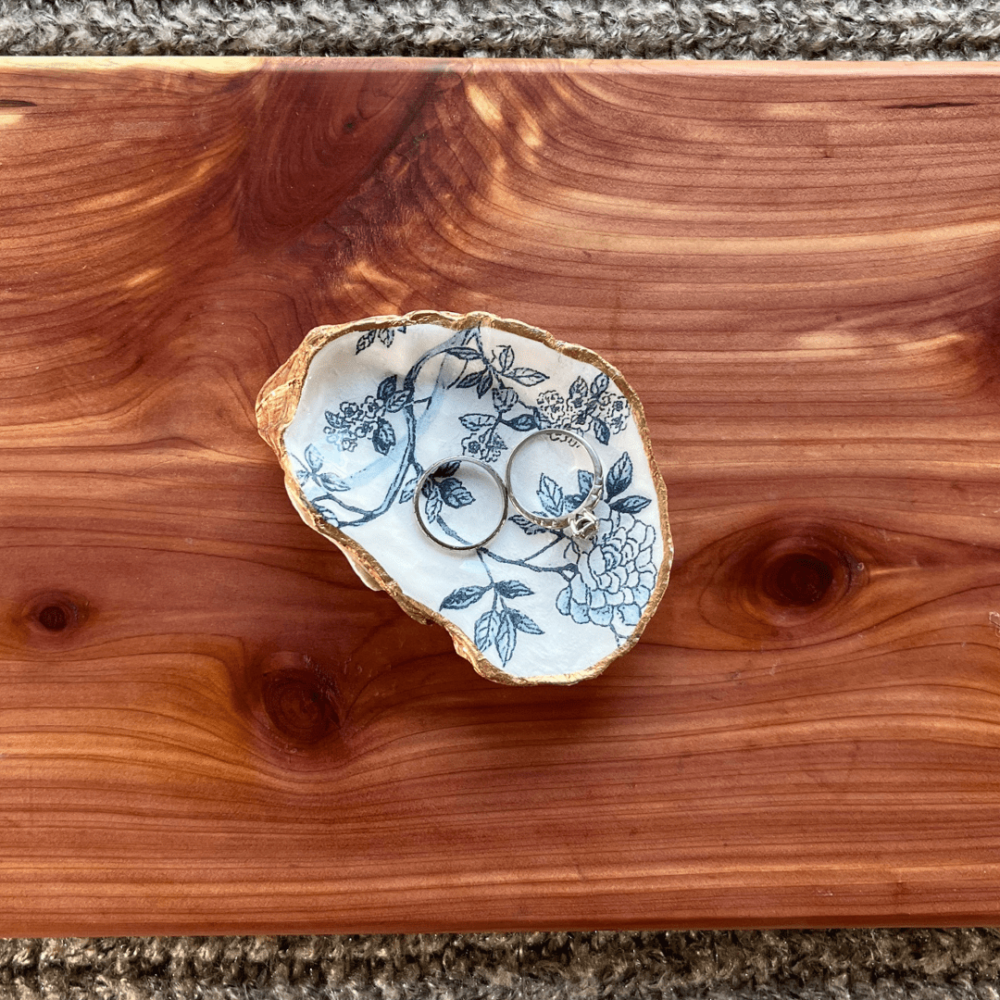 Flatlay photo of a handcrafted oyster shell ring dish with two rings in it, sitting on a pretty cedar board