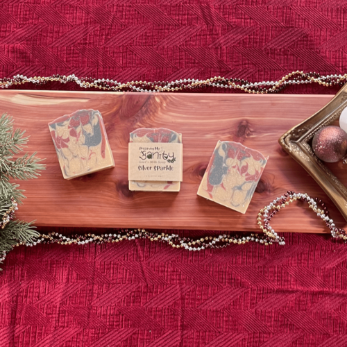 Holiday-themed flatlay of three bars of Silver Sparkle handcrafted goat's milk soap sitting on a pretty cedar bath board with a dark red backdrop