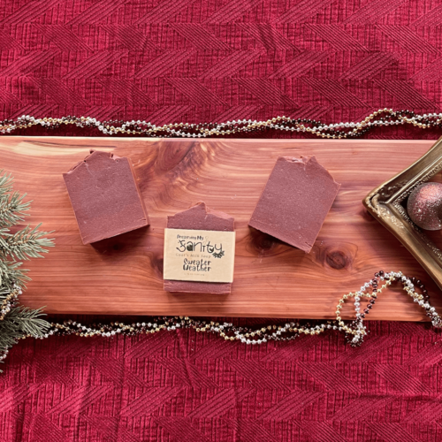 Holiday-themed flatlay photo of three bars of handcrafted goat's milk soap, Sweater Weather. Sitting on a cedar board with a dark red backdrop