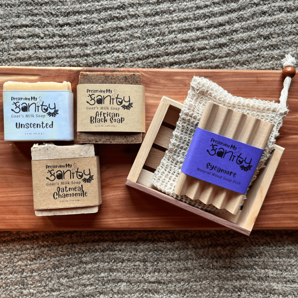 Flatlay photo showcasing the items in the unscented soap bundle, including an unscented goat's milk soap, African black soap, oatmeal chamomile soap, a wood soap dish, sisal soap saver bag, and reclaimed wood gift tray