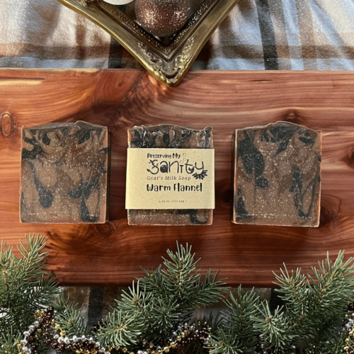 Three bars of Warm Flannel goat's milk soap on a cedar board with a holiday flatlay on a flannel shirt.