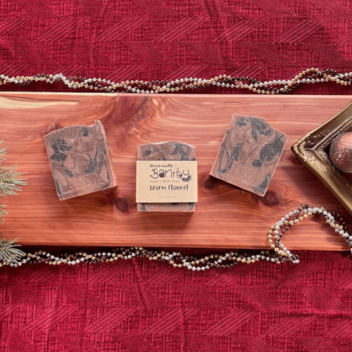 Holiday-themed flatlay of three bars of handcrafted goat's milk soap in "Warm Flannel" scent. Sitting on a cedar board with a dark red background.