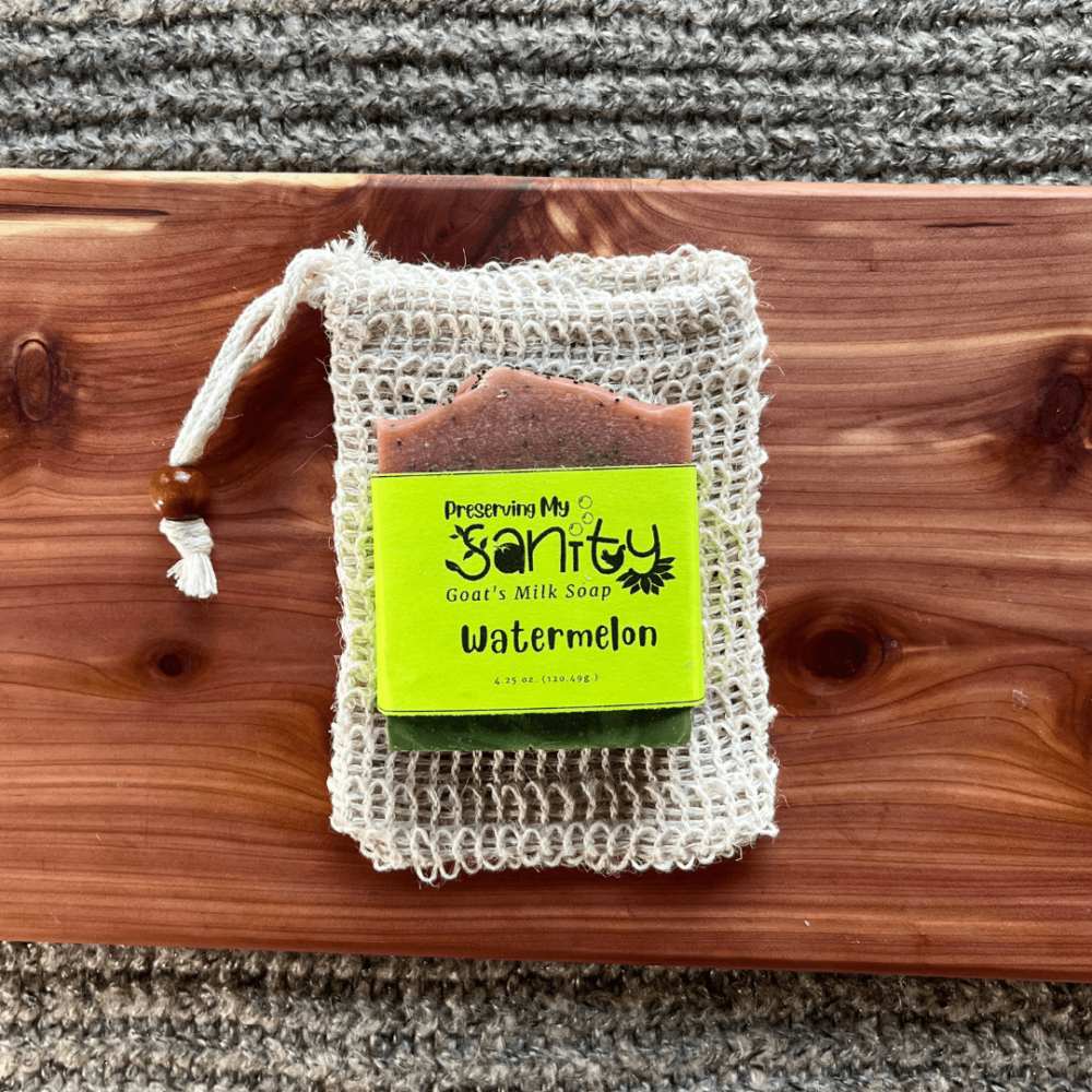 Watermelon soap stocking stuffer with a sisal soap bag, sitting on a cedar bath board with gray sweater backdrop