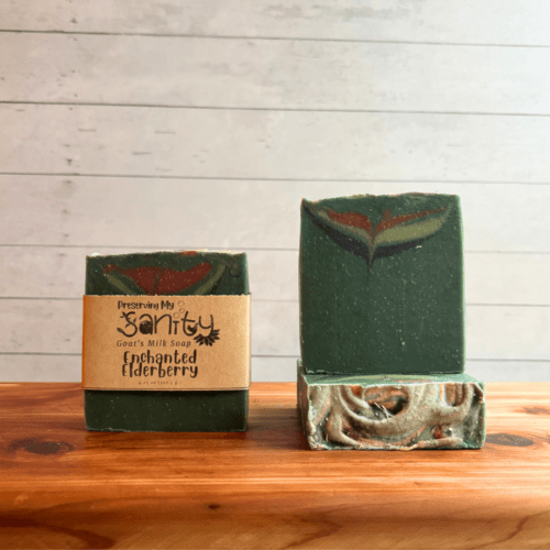 Three bars of Enchanted Elderberry goat milk soap with a white backdrop on a cedar board. Soap is dark green with a subtle blue, light green, and dark red swirl.