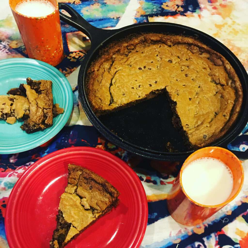 Photo of a chocolate chip cookie in a cast iron skillet with two glasses of milk and two cookies on plates.