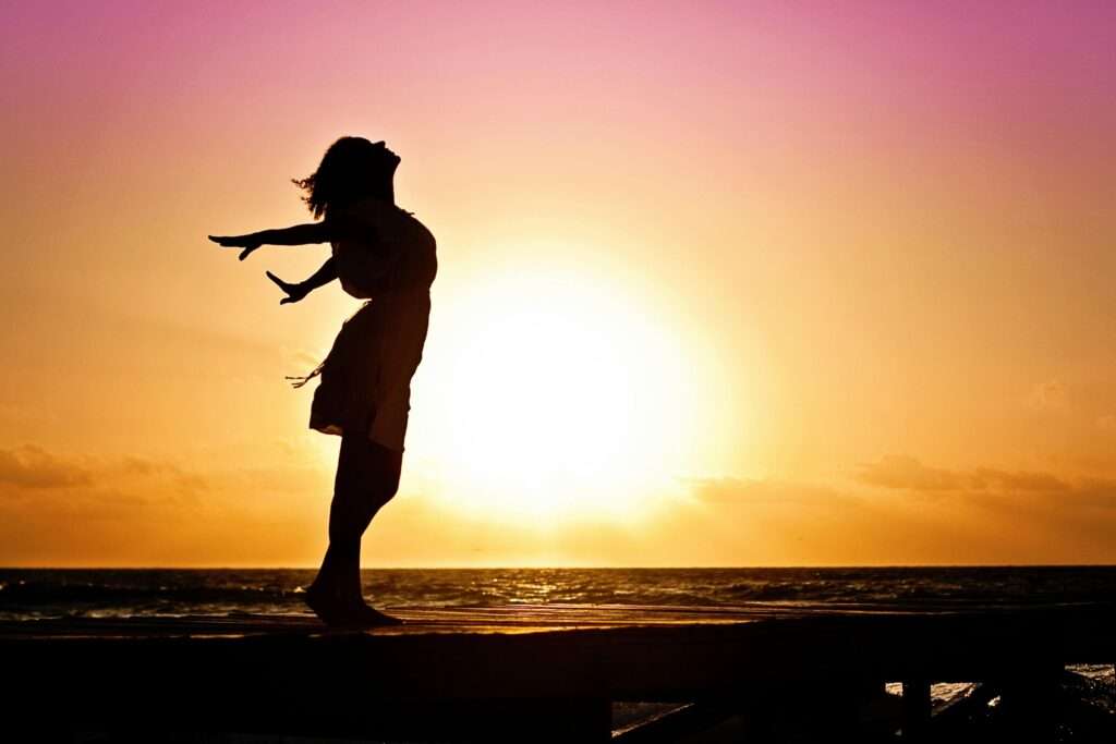 Photo of a woman standing on a beach at sunrise with her arms stretched behind her as she enjoys the beauty around her