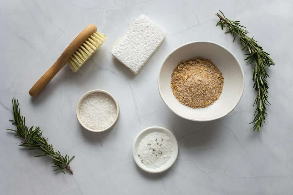 Flatlay photo of some natural products including some small bowls of salts, a brush, a sponge, and some fresh rosemary