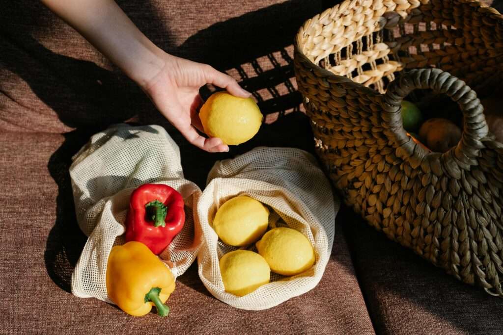 Photo of fresh produce in reusable mesh bags next to a pretty wicker basket, with a woman's hand holding a lemon also in the shot.