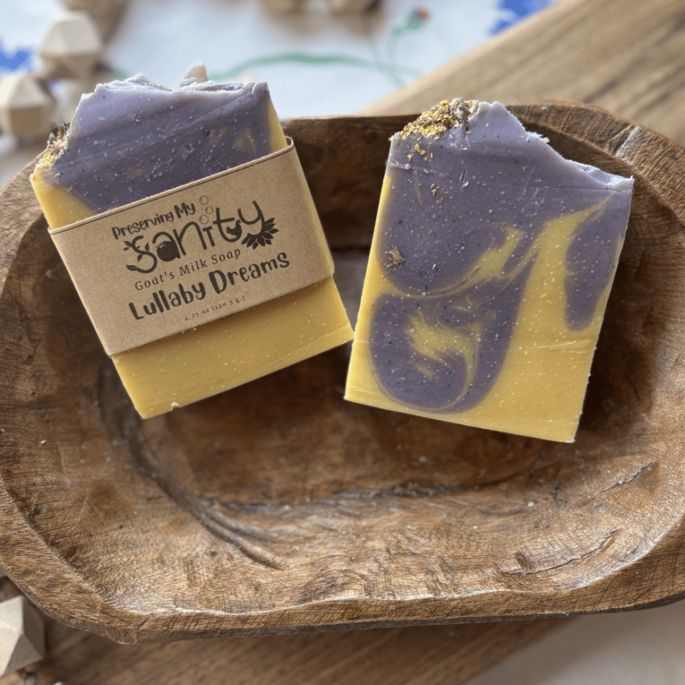 Flatlay photo of two bars of Lullaby Dreams goat's milk soap. Bars are a pale yellow color with purple drop swirl, topped with lavender and chamomile flowers.