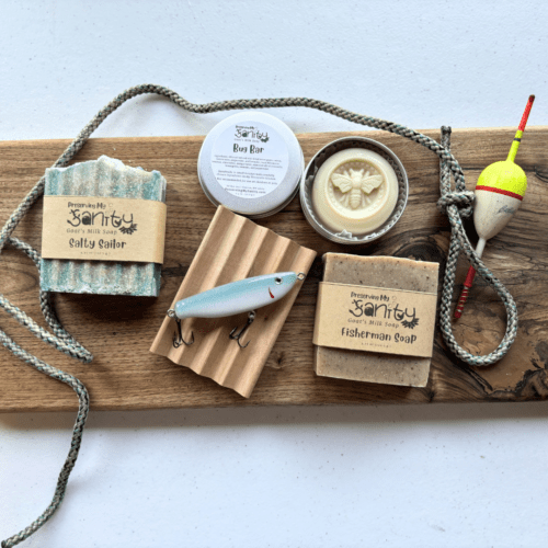 Overhead flatlay view of two bars of soap, a natural wood soap dish, a bug lotion bar in an aluminum tin, a handmade fishing lure, and some decorative props that include an old bobber, a piece of rope, and a wooden decorative board.