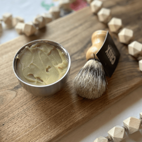 Flatlay view of a small bar of shaving soap in a stainless steel bowl with a natural wood shaving brush