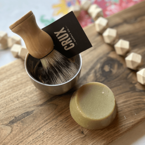 Alternate flatlay view of a small bar of shaving soap in a stainless steel bowl with a natural wood shaving brush