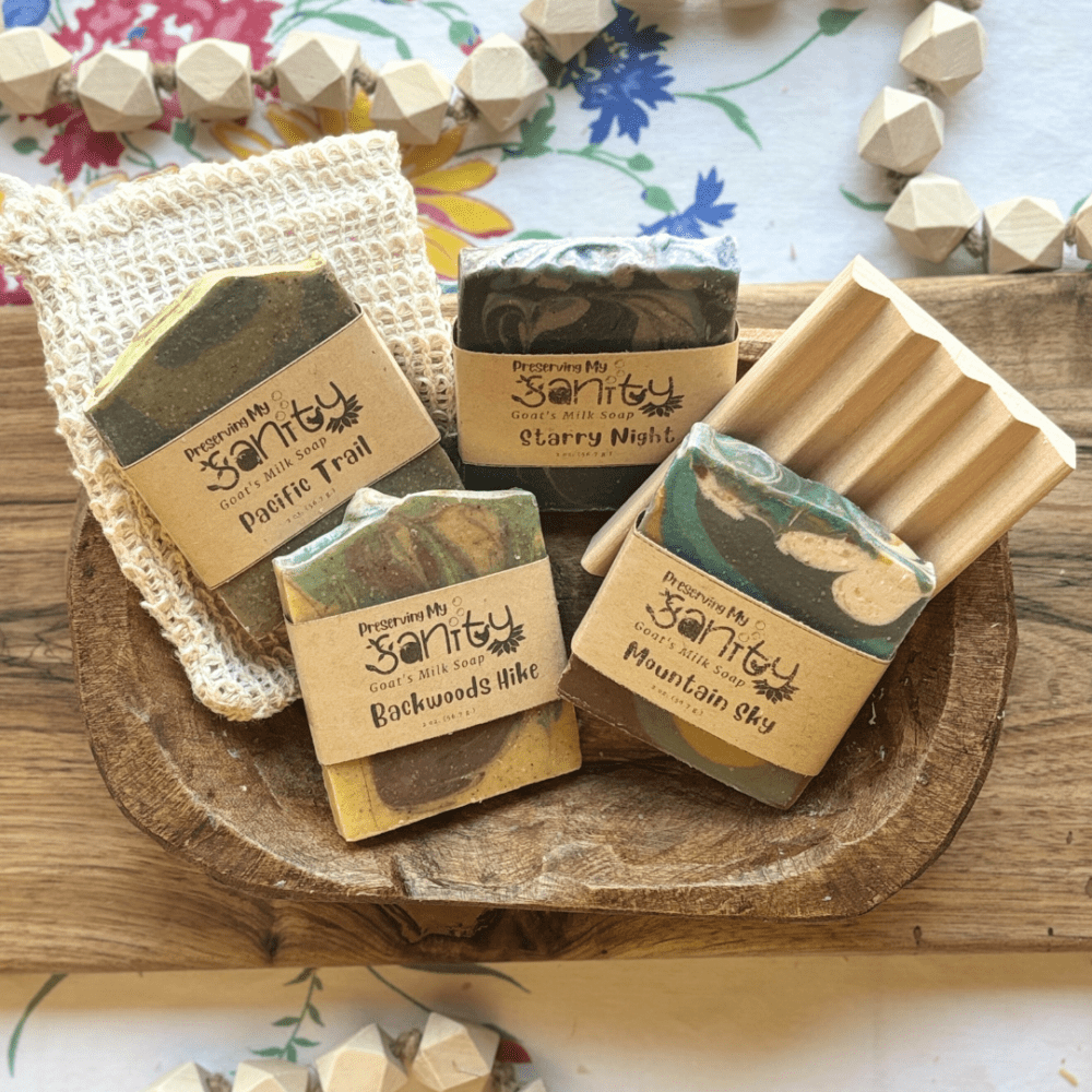 Overhead flatlay view of the masculine soap sampler, featuring four half-size bars of soap, a soap saver bag, and a natural wood soap dish - all sitting in a decorative wooden bowl on a flatlay display of a board, tablecloth, and wooden beads