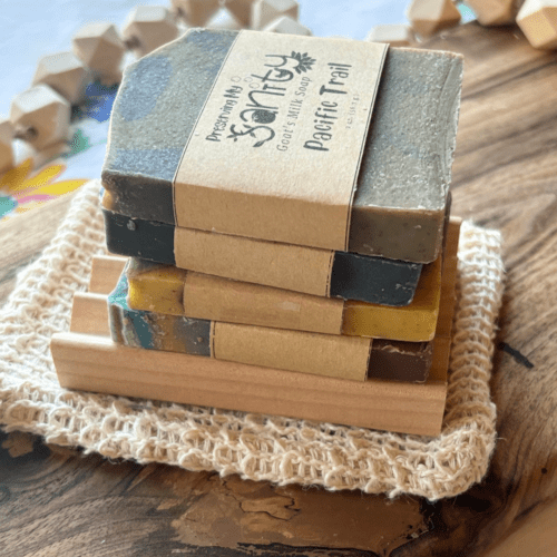 Side view of the items included in the masculine soap sampler - four half-bars of goat's milk soap, a natural wood soap dish, and a soap saver bag - all sitting on a wood board.