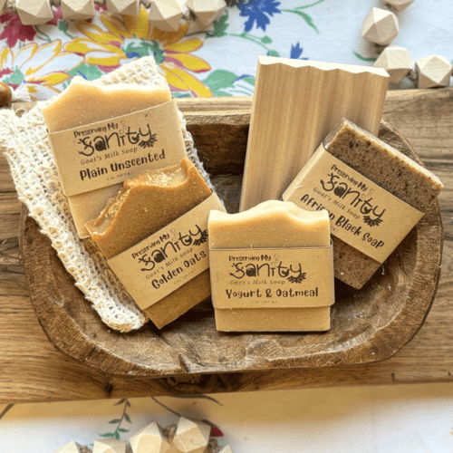 Overhead flatlay view photo of four half-size bars of unscented soap, a natural wood soap dish, and a natural fiber soap saver bag sitting in a decorative wooden bowl