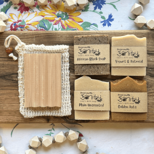 Overhead flatlay view photo of four half-size bars of unscented soap, a natural wood soap dish, and a natural fiber soap saver bag laying on a handmade wood board