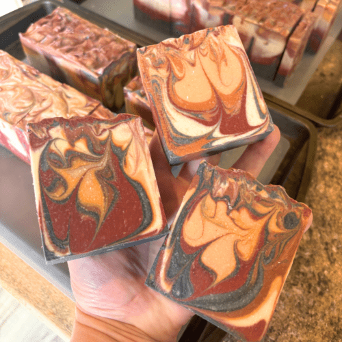 Photo showing process of goat milk soap with three bars held in maker's hand just after cutting soap loaves