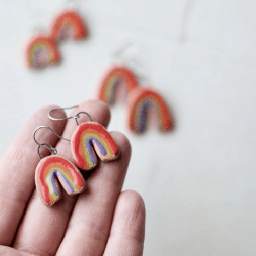 Photo showing an outstretched hand holding one pair of ceramic rainbow earrings, with two other pairs blurred out amid a white background