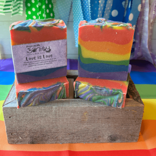 Photo of colorful 6-layer stripe goat's milk soap to celebrate Pride Month. The name of the soap is "Love is Love" and the photo features four bars displayed at different angles with a colorful backdrop.