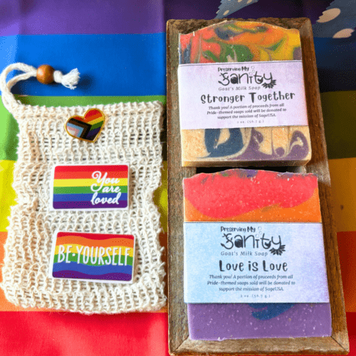 Flatlay photo showing items included in the Pride soap sampler - two half bars of Pride Month soap, a sisal fiber soap saver bag, two stickers, and a lapel pin